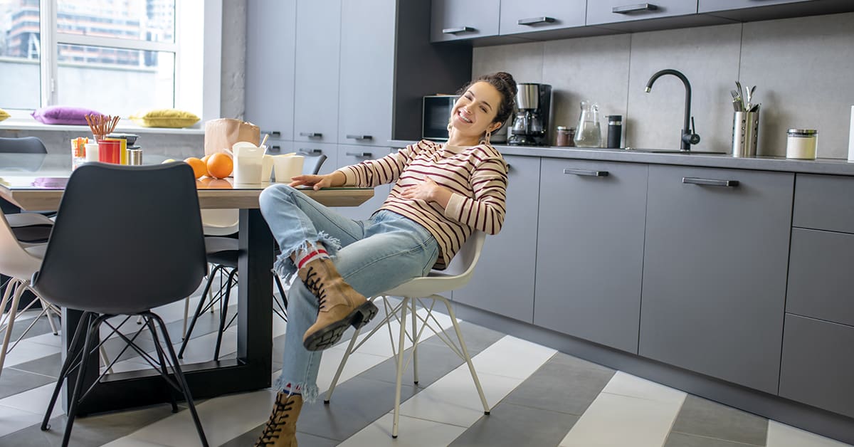 Indoors. Young woman sitting at the kitchen table with crossed leg, holding stomach, smiling