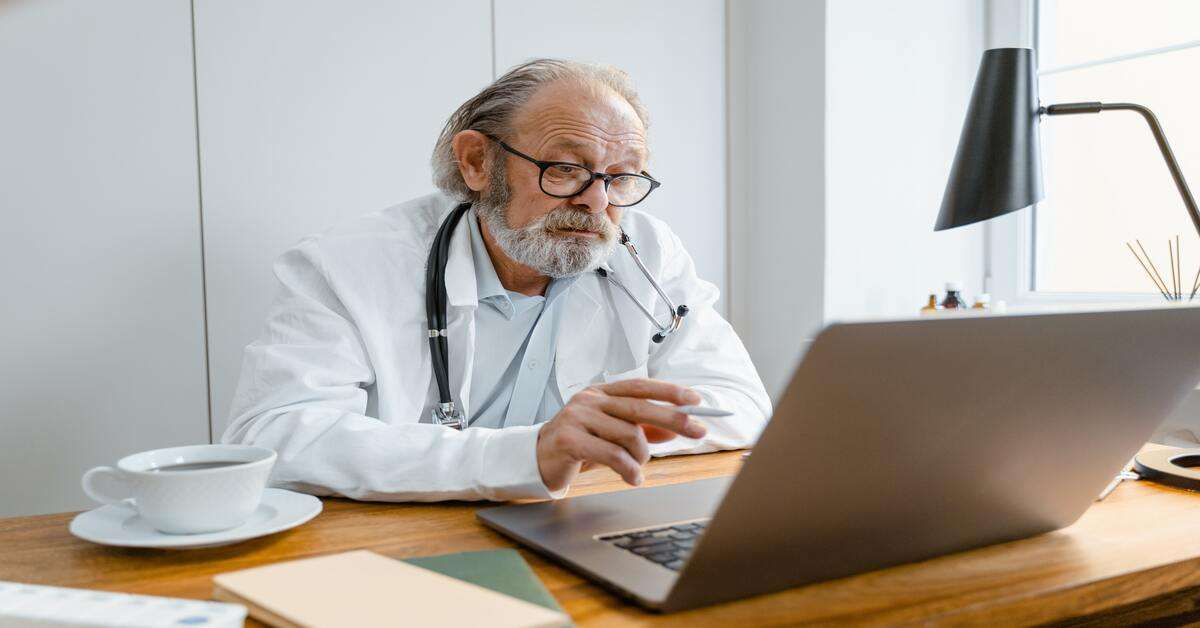 A senior doctor works at a laptop.