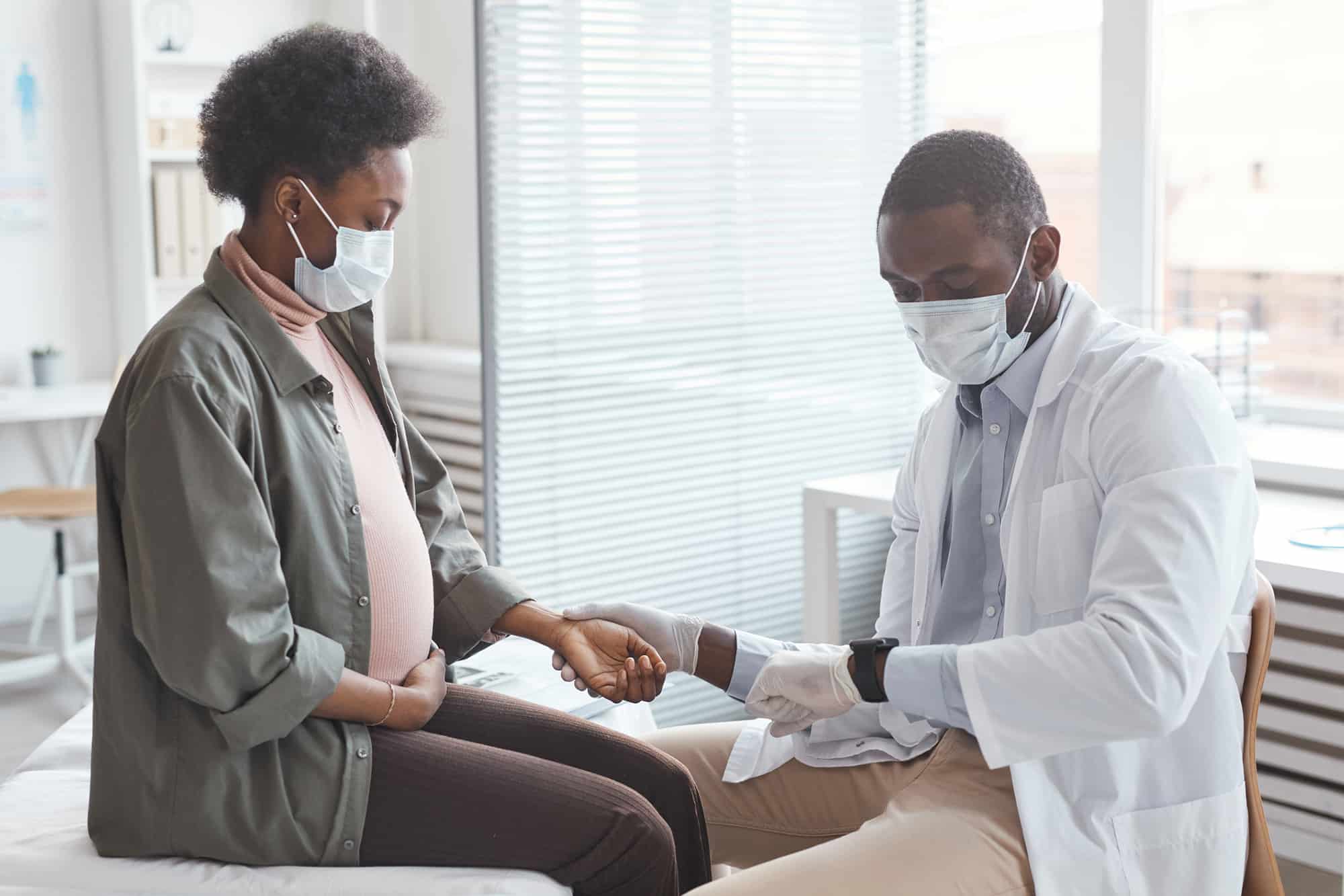 African young man in mask measuring the pulse of pregnant woman and caring about her health at hospital