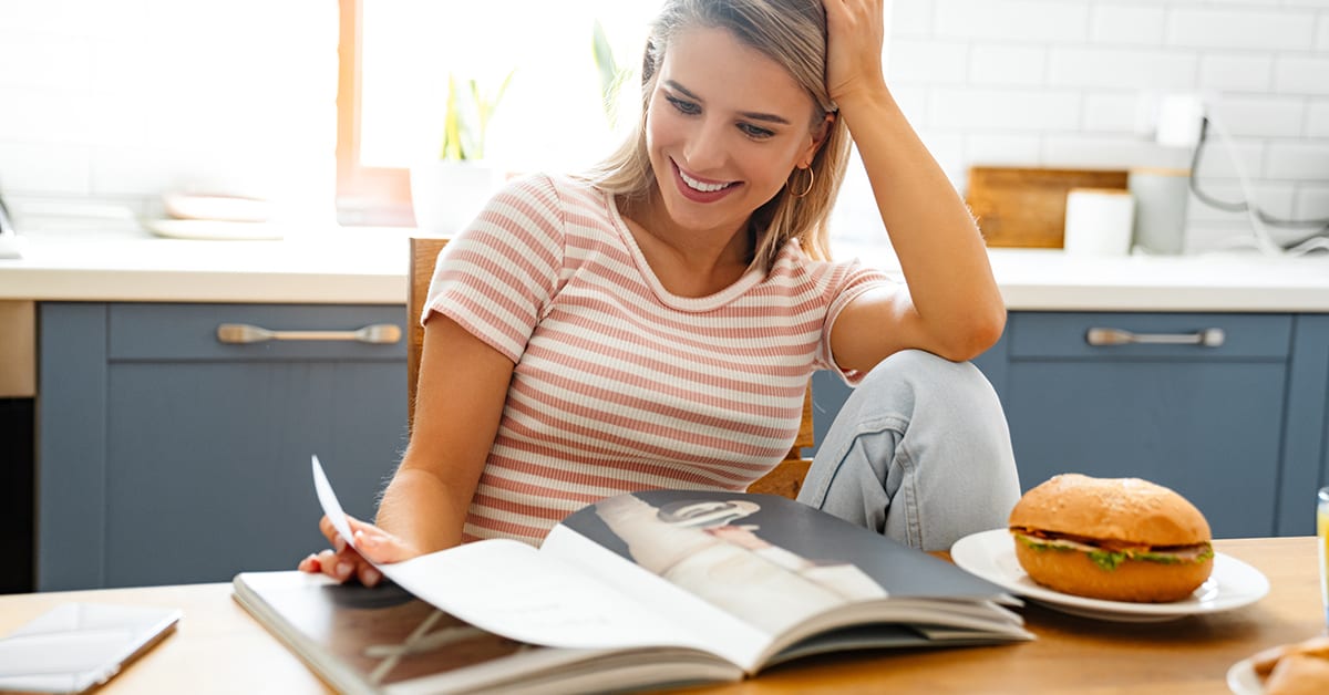 Smiling young woman eating a hamburger at home, looking through magazine