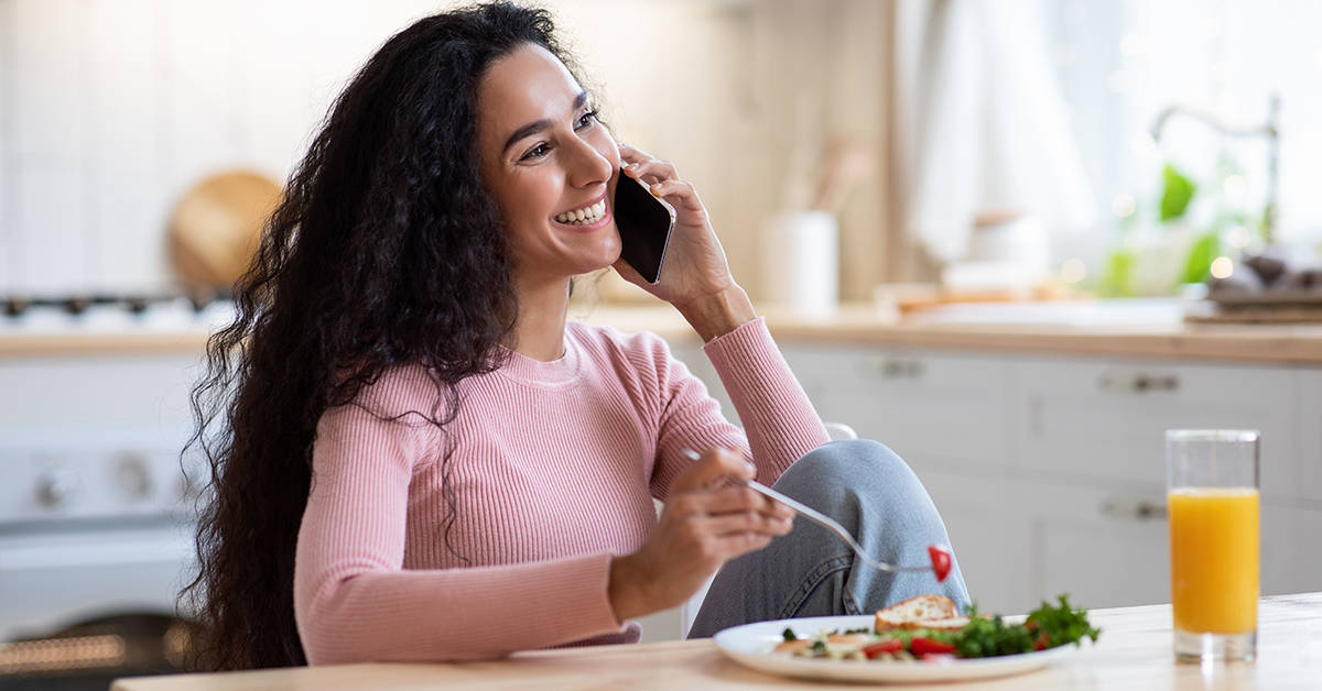 Smiling Millennial Lady Eating Breakfast And Talking On Phone In Kitchen At Home, Cheerful Young Brunette Woman Enjoying Tasty Food While Having Pleasant Mobile Conversation